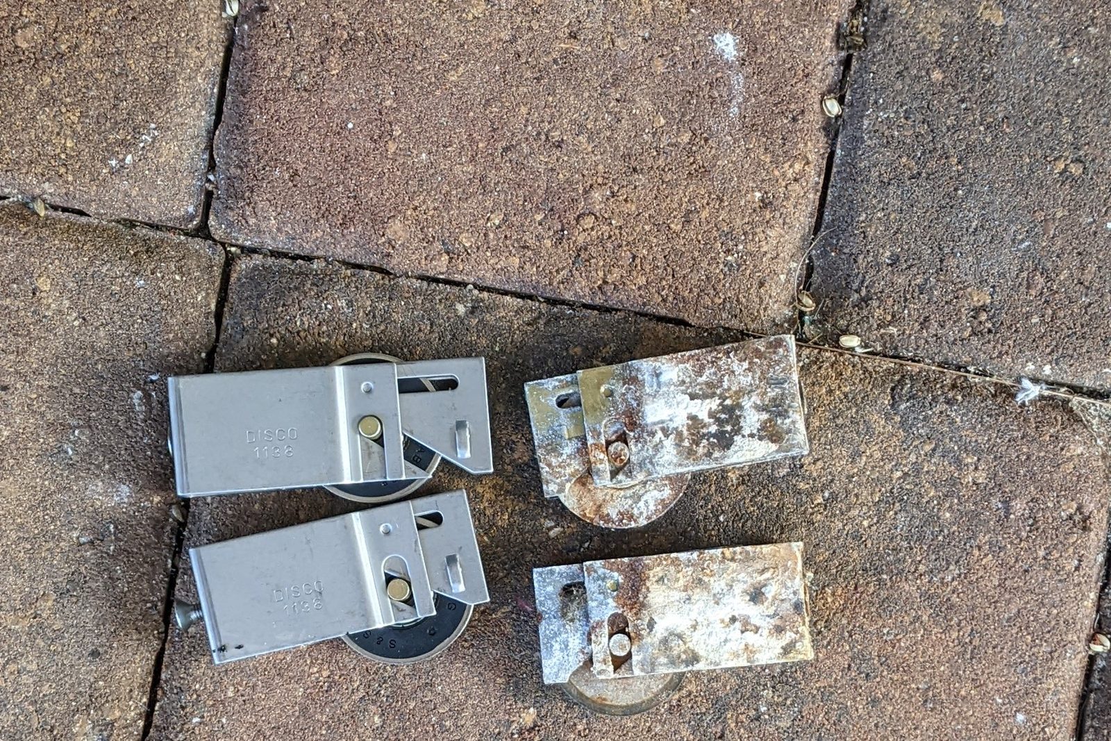Two corroded sliding door rollers beside two new replacement rollers on a paver patio